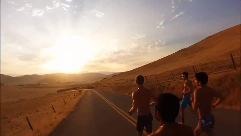 Five men are running shirtless on a paved road through a sunlit, hilly landscape. The sun is either rising or setting, casting a warm glow over the scene. The terrain is dry with scattered fencing along the road, and mountains are visible in the distance.