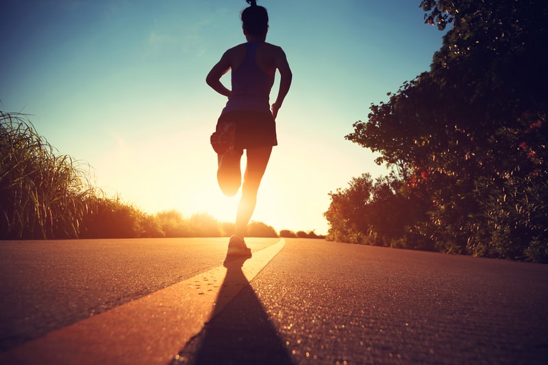 A person jogs on a paved road at sunrise. They are shown from the back, with shadows cast on the ground. The surrounding area includes lush greenery, and the sky is lit with the golden hues of the early morning.