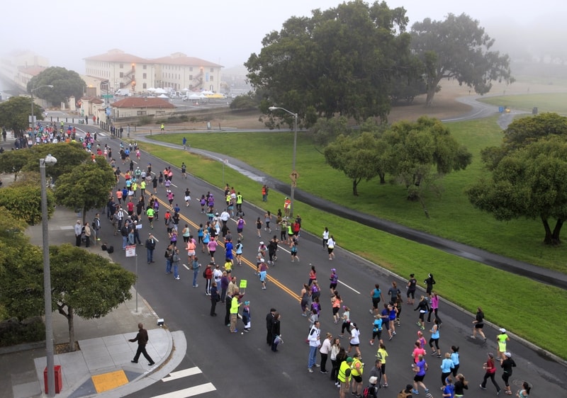 A large crowd of runners participating in a race on a street bordered by green lawns and trees. Residential buildings and parked cars are visible in the background under an overcast sky with fog. Spectators and volunteers stand along the street.