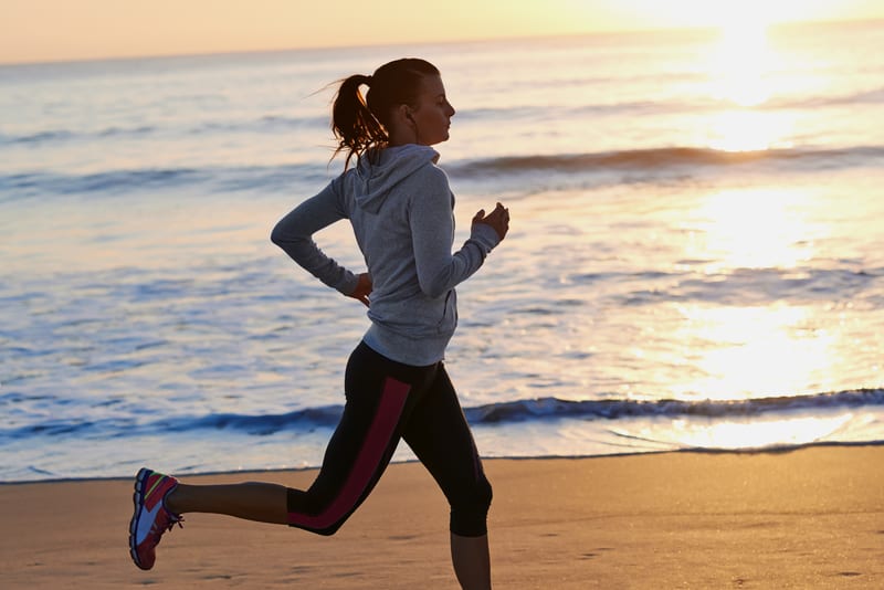 A person wearing athletic gear, including a gray hoodie and black leggings with red stripes, jogs along a beach at sunrise. The sun is low on the horizon, casting a golden glow over the ocean and creating a serene, peaceful atmosphere.