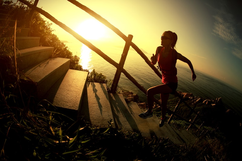 A silhouette of a woman running up outdoor stairs along a seaside path, with a wooden handrail. The sun is setting, creating vibrant golden hues in the sky and reflecting on the water. The scene evokes a sense of determination and tranquility.