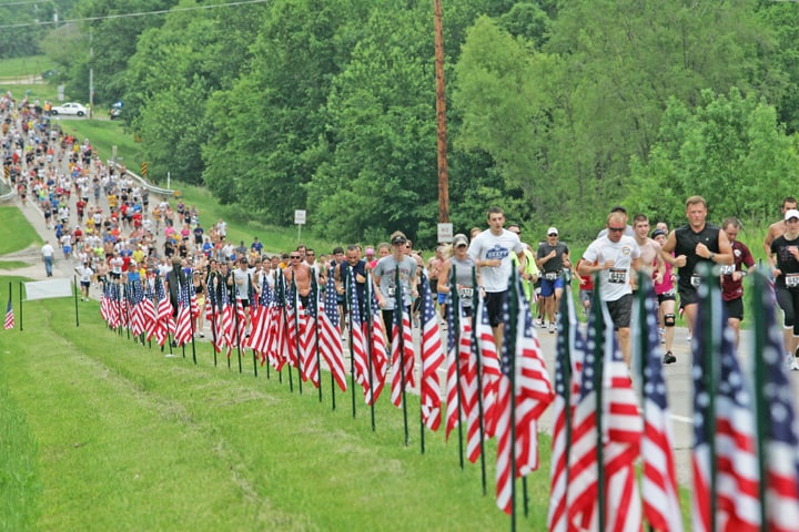 A large group of runners participating in a race on a road lined with numerous American flags on both sides. The scene is set in a lush, green, forested area on a cloudy day, giving the photo a vibrant yet serene backdrop.