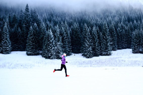 A person dressed in a purple jacket and black pants runs across a snow-covered landscape with a dense forest of snow-dusted evergreen trees in the background. Thick fog hovers above the treetops, creating a cold and serene atmosphere.