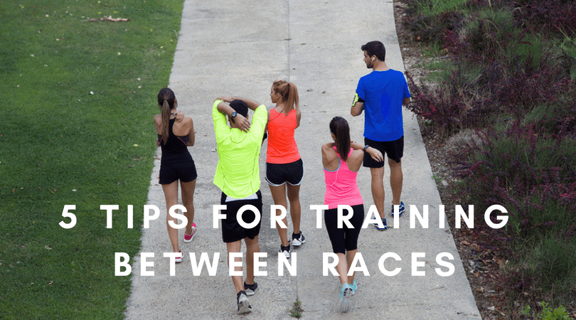 Overhead view of five people in athletic wear walking on a path surrounded by grass and trees. Text overlay reads, "5 TIPS FOR TRAINING BETWEEN RACES.