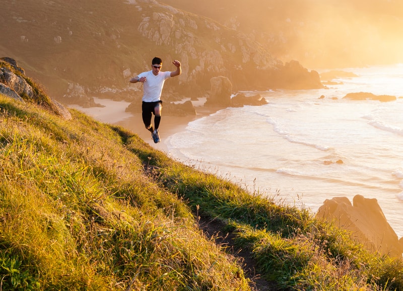 A person in athletic clothing runs energetically along a grassy, sunlit hillside trail near the ocean. The sun sets in the background, casting a warm golden glow over the landscape, with waves crashing against the rocky shore below.