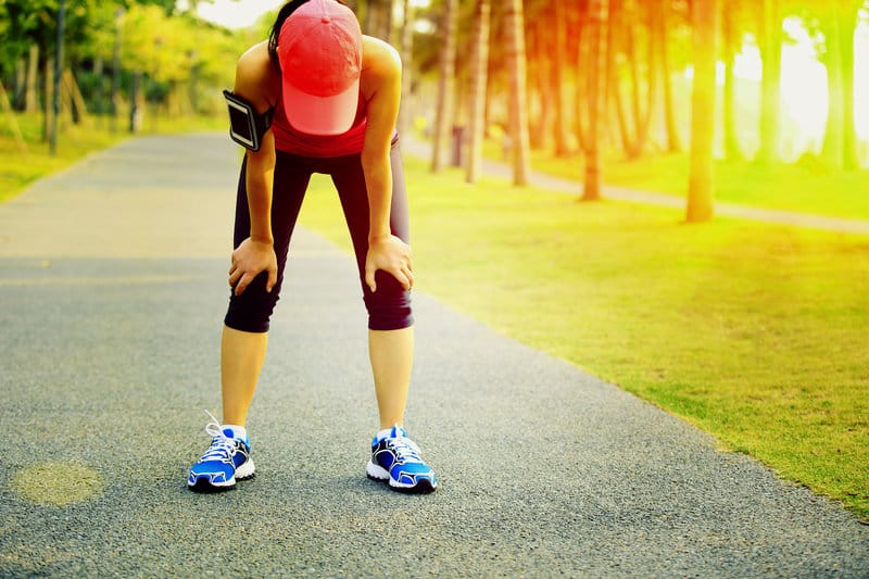 A person wearing a pink cap, black athletic wear, and blue running shoes is bent over with hands on knees, appearing tired or out of breath. They are on a paved path with sunlight filtering through trees in the background.