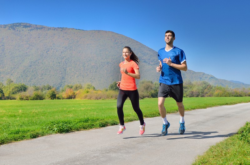 A man and a woman are jogging on a paved path in an open field with a backdrop of green mountains under a clear blue sky. The woman wears a red T-shirt and black leggings, while the man wears a blue T-shirt and black shorts. They appear to be enjoying their run.