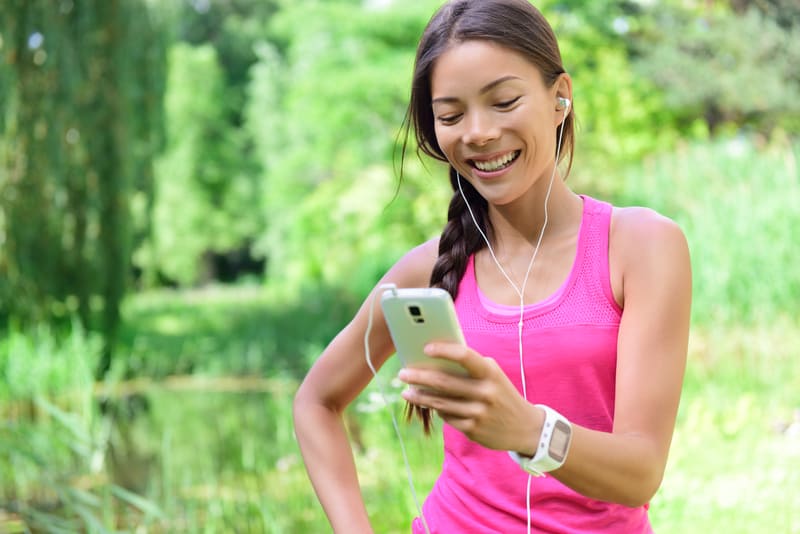 A woman in a pink tank top is outside in a green, natural setting, smiling and looking at her smartphone. She has earphones on and a smartwatch on her wrist, suggesting she might be listening to music or tracking her exercise.