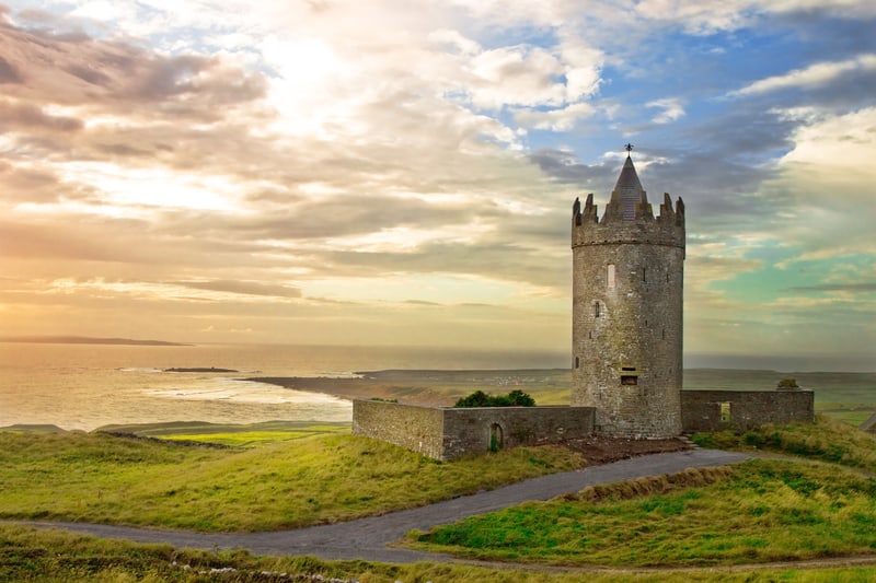 View of Doonagore Castle, a tower house on a hill overlooking the Atlantic Ocean in County Clare, Ireland. The castle is set against a background of a colorful sky with scattered clouds and the landscape features green fields and a curving road leading to the tower.