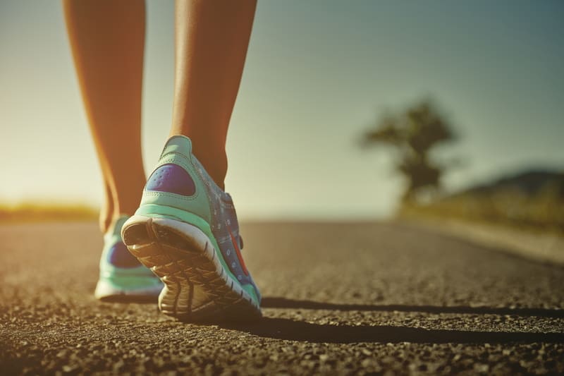 Close-up of a person's legs and feet wearing turquoise and purple running shoes, walking on an asphalt road with blurred background of scenery and a tree under a clear sky. The image is taken from behind and focuses on the footwear and lower legs.