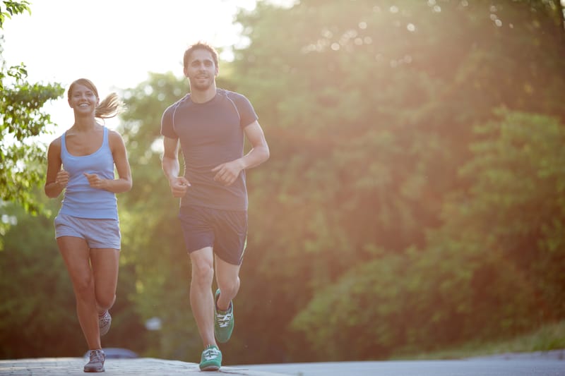 A woman and a man are jogging at sunrise or sunset on a paved path with trees in the background. Both are smiling; she is wearing a light blue tank top and shorts, and he is in a black T-shirt and shorts. The scene is bathed in warm, natural light.