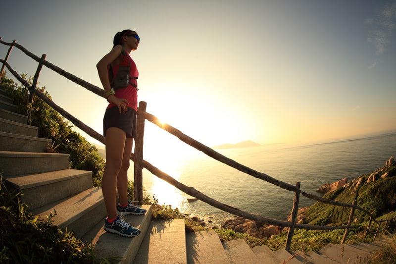 A person in athletic gear and sunglasses stands on stone steps by a coastal path, gazing at the ocean during a sunrise or sunset. They are surrounded by green foliage and a wooden railing, with the sun casting a warm glow over the scene.