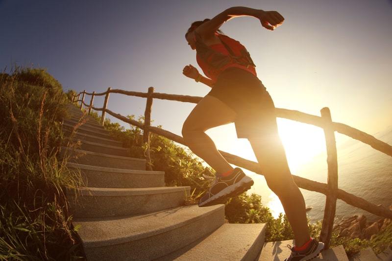 A person in athletic wear runs up a set of outdoor stairs surrounded by wooden railings and greenery. The sun is low in the sky, casting a warm glow and long shadows, suggesting either early morning or late afternoon. The scene has an energetic and motivating feel.