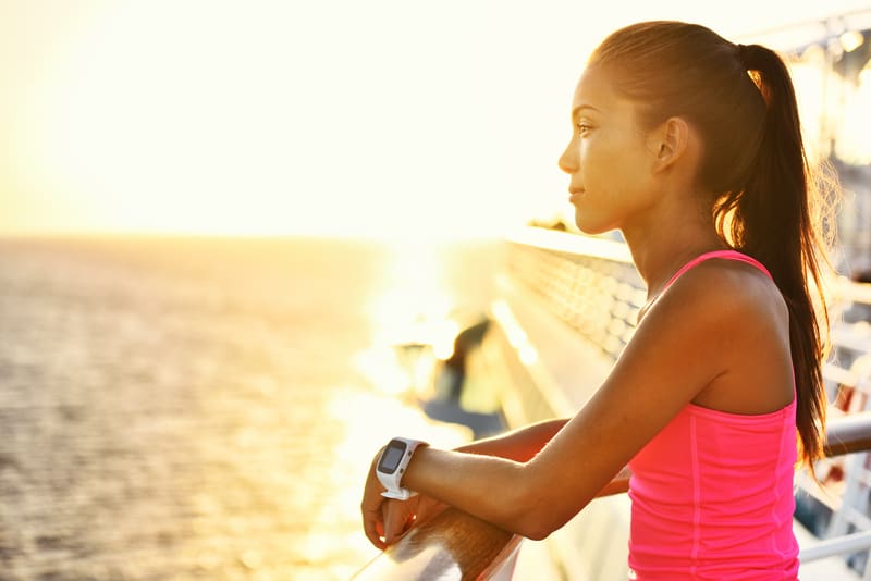 A woman wearing a bright pink tank top leans on a railing, gazing out at the ocean during sunset. She has her hair tied back and is wearing a smartwatch on her wrist. The sunlight casts a warm, golden glow on the scene.