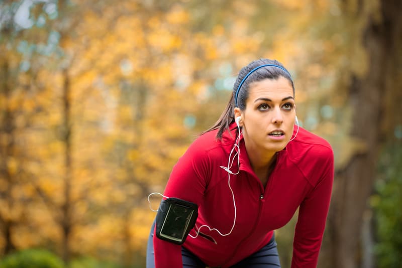 A woman in a red jacket, wearing earphones, pauses and leans forward while jogging in a scenic park during autumn. She has a smartphone strapped to her arm and looks focused.