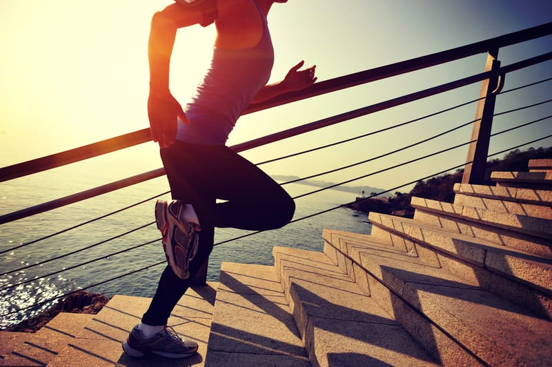 A person runs up outdoor stairs near a body of water during sunset. The sun casts a warm golden light, creating elongated shadows. The individual, wearing athletic clothing and sneakers, appears determined and energetic against the scenic backdrop.