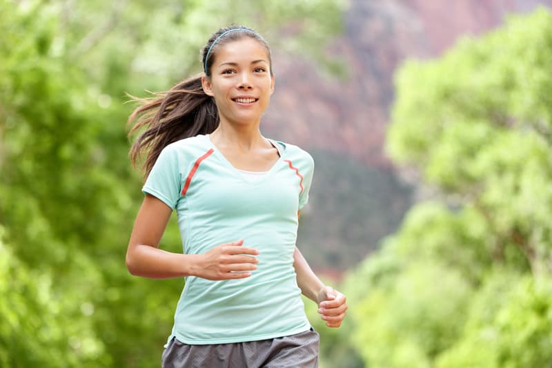 A woman wearing a light blue shirt and gray shorts is jogging outside. She has a ponytail and a headband. She appears to be running on a trail surrounded by greenery, with trees and blurred natural scenery in the background.