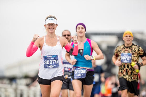 A group of runners participating in a race. The leading female runner wears a white tank top, pink arm sleeves, a white visor, and a race bib numbered 30070. The runners behind her wear colorful attire and visible race numbers. Buildings can be seen in the background.