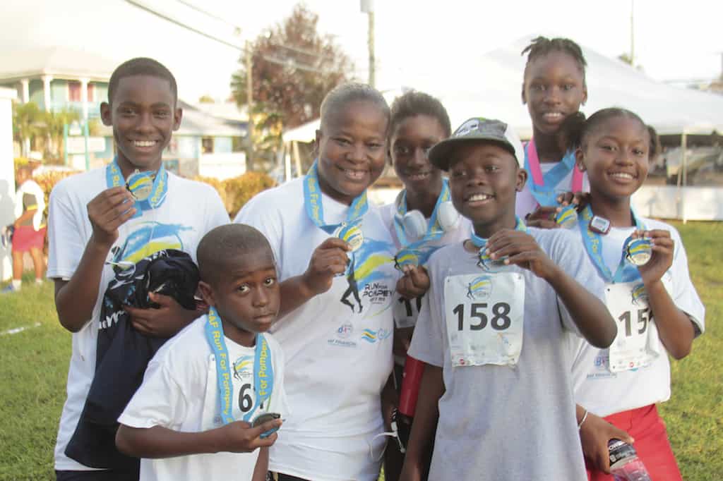 A group of smiling individuals, including children and adults, pose together outdoors holding medals and wearing race bibs. They appear to have just completed a race or fun run, as indicated by their athletic gear and joyful expressions.