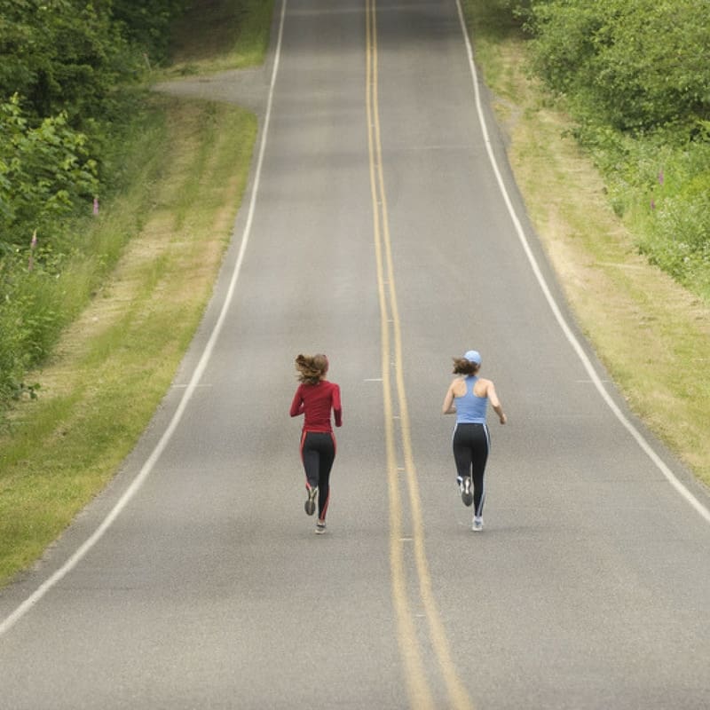 Two people are running on an empty road flanked by lush greenery. The road stretches far into the distance. One wears a red top and black pants, while the other wears a light blue top with a white cap. The scene is calm and serene.