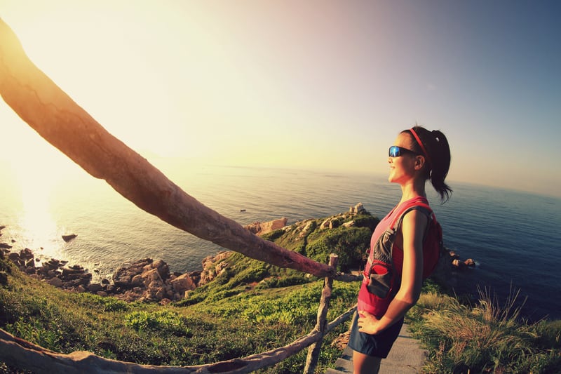 A woman wearing sunglasses and a backpack stands on a pathway overlooking a scenic ocean view at sunset. She smiles, enjoying the moment, as the sun casts a warm glow on the landscape, with rocky cliffs and green vegetation in the foreground.