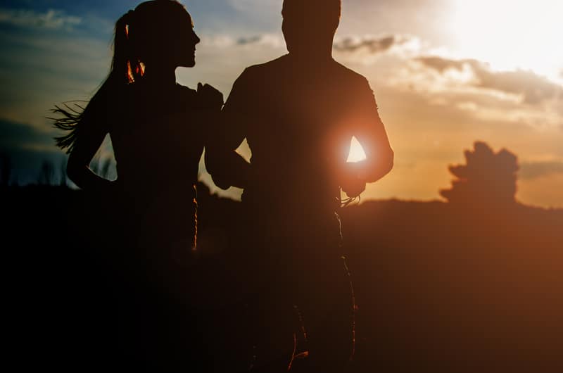 Silhouetted image of two people jogging together at sunset. The sky is vibrant with shades of orange, yellow, and blue, while the sun casts a warm glow, creating a serene and picturesque scene.