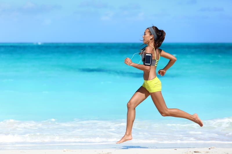 A woman in athletic wear jogs along the shore of a pristine beach with clear turquoise water under a blue sky. She has headphones on and a smartphone strapped to her upper arm.