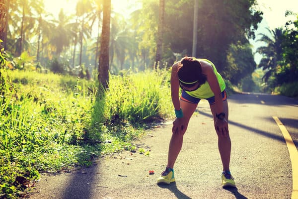 A person in athletic gear, appearing exhausted, bends over with hands on knees while standing on a sunny road surrounded by greenery and palm trees. The sun shines through the trees, casting a warm glow over the scene.