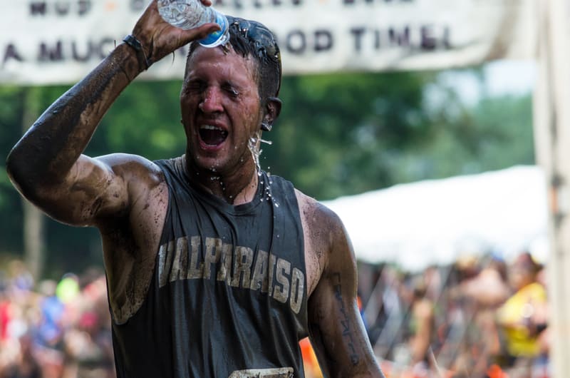 A man, covered in mud and sweating, pours water over his head to cool down during an outdoor event. He is wearing a sleeveless shirt with text on the front, and the background shows blurry trees and spectators.