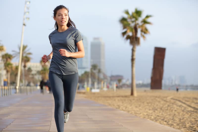 A woman jogs on a wooden pathway by the beach on a clear day. Palm trees and tall buildings are visible in the background, with the ocean to the side. She wears a grey t-shirt and black leggings, looking straight ahead as she runs.