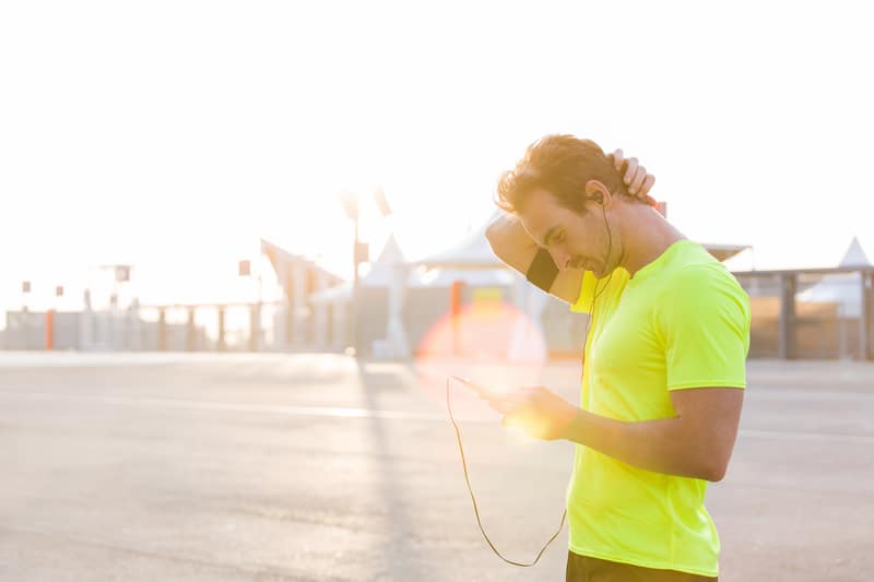 A person wearing a bright yellow t-shirt stands outdoors in a sunlit area, looking down at a device held in their hand. They are wearing earphones and have one hand behind their neck. The background features a clear sky and industrial buildings.