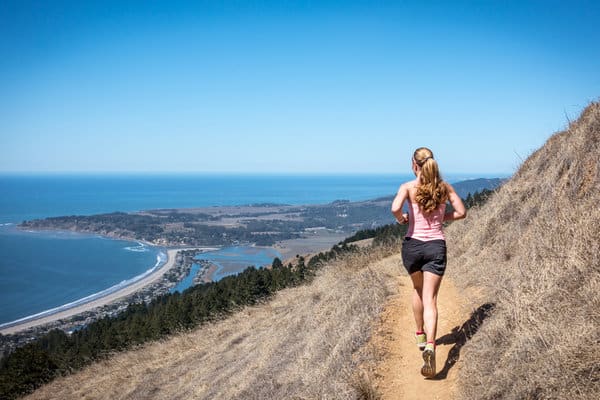 A woman with a ponytail runs on a narrow dirt trail along a hillside overlooking an expansive coastal landscape with a clear blue sky, the ocean, and a stretch of sandy beach in the distance. She wears a pink tank top, black shorts, and running shoes.