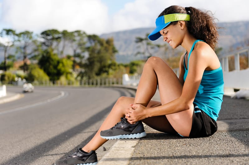 A woman in athletic attire sits on the curb of an empty road, holding her ankle with a look of discomfort on her face. She wears a blue tank top, black shorts, and a blue visor. In the background, trees and mountains are visible under a clear sky.