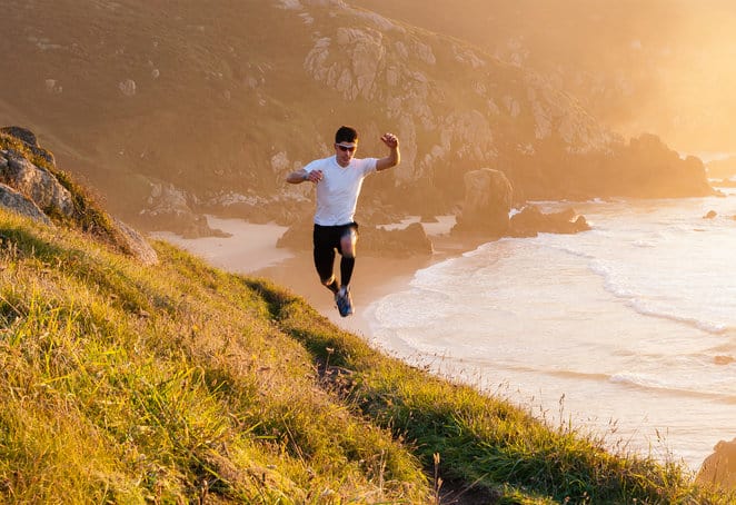 A person wearing a white shirt and black shorts is running energetically down a grassy hill near a coastline at sunset. The ocean waves crash against the rocks below, and the scene is bathed in a warm, golden light.