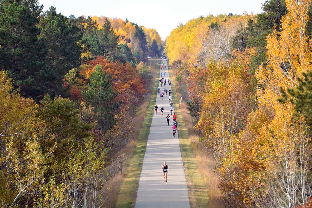 An aerial view of a marathon taking place on a paved path through a forest during autumn. Runners are seen spread out along the long, straight path, surrounded by trees in brilliant fall colors of yellow, orange, and red.