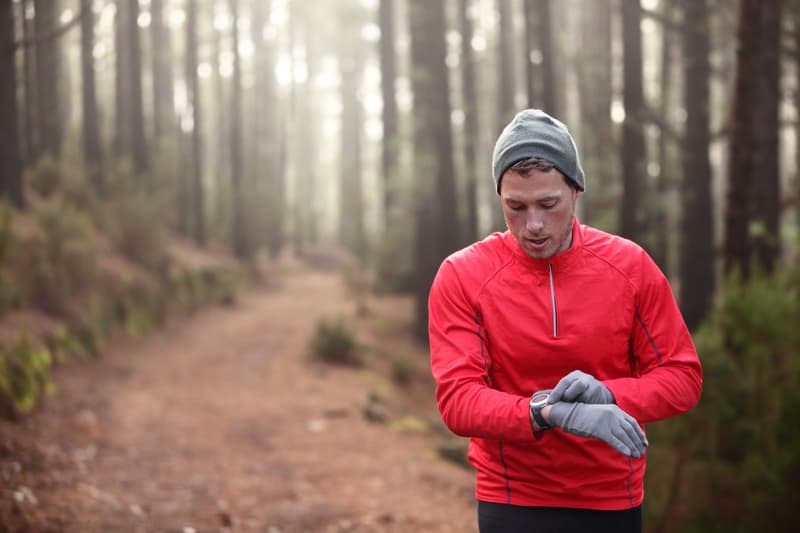 A person wearing a red jacket, grey gloves, and a grey beanie is checking their smartwatch while standing on a forest trail. The background includes tall trees and a dirt path extending into the distance.