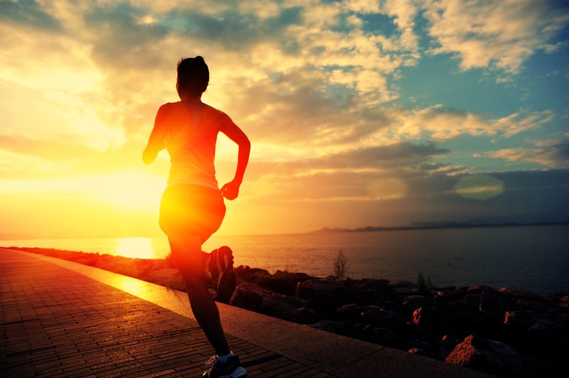 Silhouette of a person running along a waterfront path during a vibrant sunset. The sun is low in the sky, casting a warm glow over the scene, while clouds are scattered across the colorful sky. The water appears calm, reflecting the hues of the sunset.