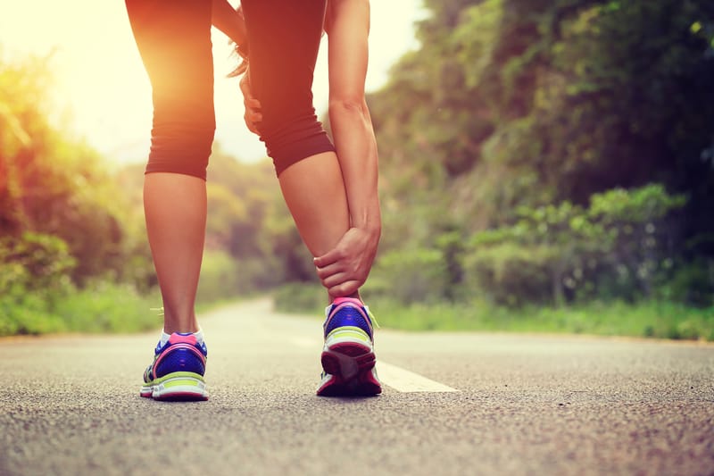 A runner standing on a paved path in the outdoors, bending down and holding their ankle as if in pain. The person is dressed in athletic wear, including bright running shoes. The surroundings are lush and green, and the sun is setting or rising in the background.