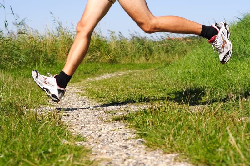 A person in running shoes and shorts is mid-stride while running on a gravel path surrounded by tall grass on a sunny day. Only their legs are visible in the image.