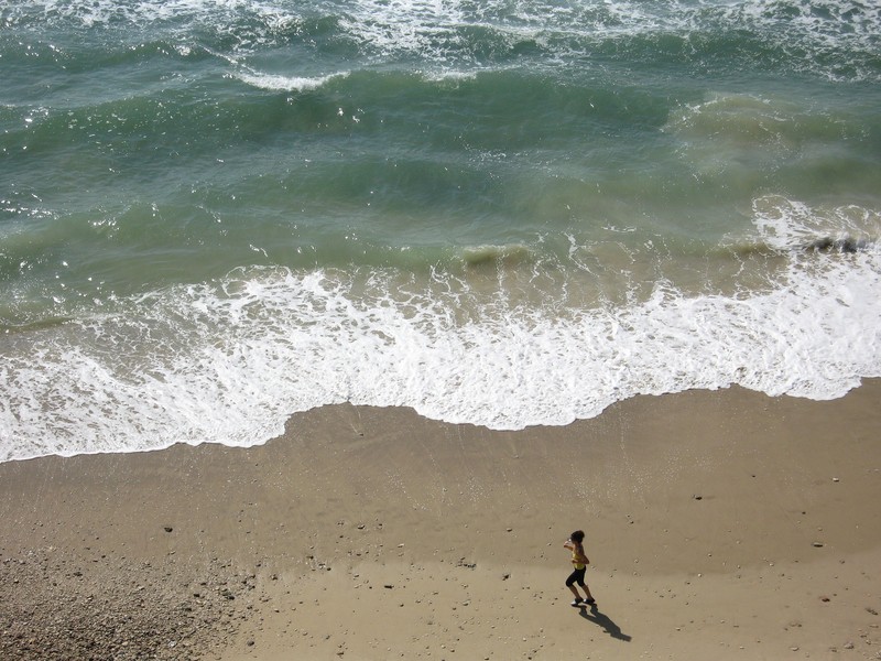 A person runs along a sandy beach near the shoreline, where foamy waves gently meet the sand. The ocean stretches out into the distance, with shades of blue and green waters. The scene is sunny with clear skies, and the beach appears mostly empty.