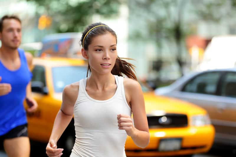 A young woman in a white tank top is jogging on a city street, with a yellow taxi and a man in a blue tank top running behind her. The background features blurred urban scenery.