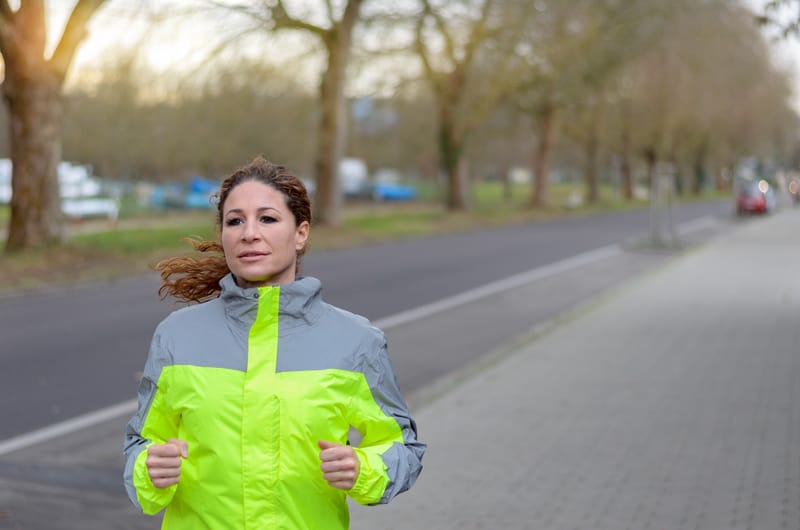 A woman is running along a tree-lined road wearing a fluorescent yellow and gray jacket. The background features a pathway, bare trees, and out-of-focus parked cars. She appears focused on her run.