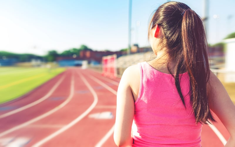 A person with long hair tied in a ponytail, wearing a pink tank top, stands on the side of an outdoor running track, facing away from the camera. The track curves ahead and there are bleachers and a field in the background.