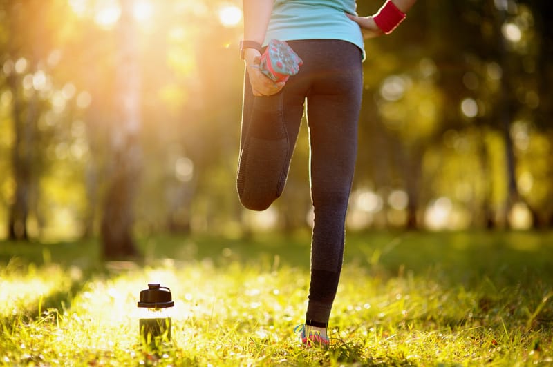 A person dressed in athletic wear stretches one leg while standing on a grassy field in a forest. The individual is holding their foot behind them with one hand. A water bottle is placed on the ground nearby. Sunlight filters through the trees in the background.