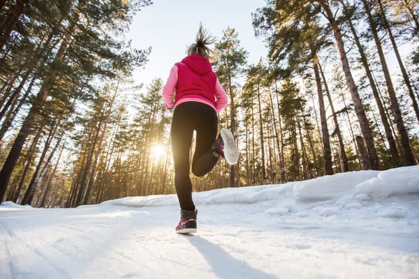 A person wearing a pink hoodie and black pants is running on a snowy path through a forest with tall trees. The sun is shining brightly through the trees, casting long shadows on the snow.