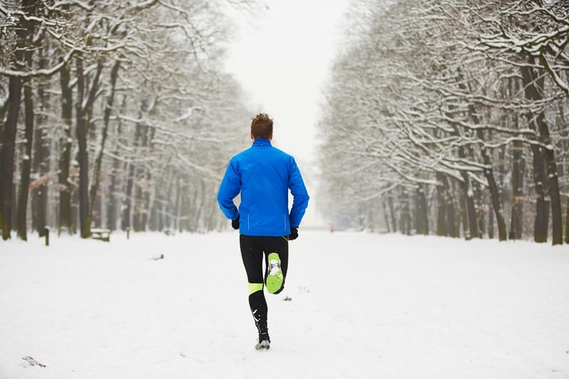 A person wearing a blue jacket and black leggings with neon green accents is jogging through a snowy, tree-lined pathway. Snow covers the ground and the trees, creating a serene, wintery atmosphere. The person is seen from behind, moving away from the camera.