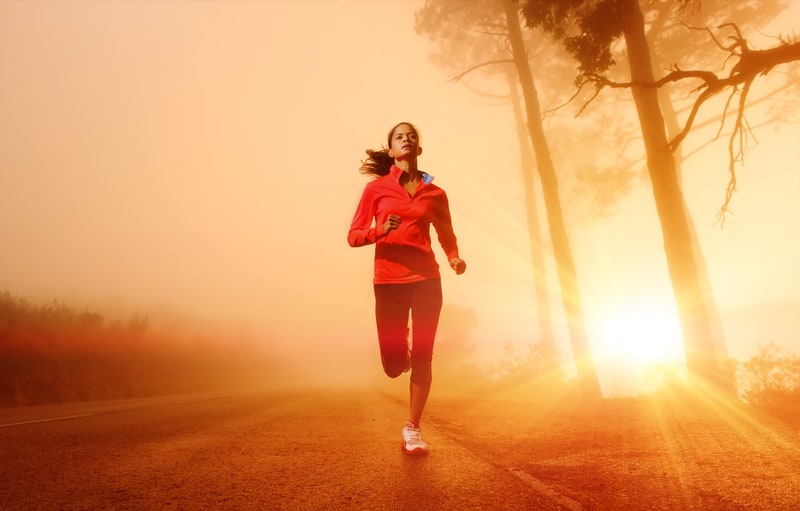 A woman in a red jacket and black leggings is running on a foggy road with tall trees on either side. The sunlight breaks through the mist, casting a warm golden glow over the scene.