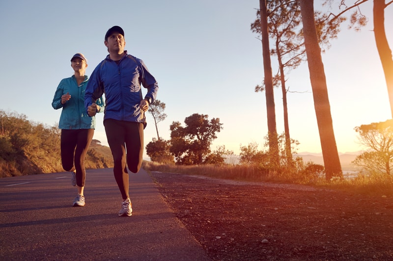 Two people are running on a paved road during sunset, with trees on the sides. They both wear running clothes, including caps and jackets. The scenery includes distant mountains and a clear sky, suggesting a peaceful, outdoor exercise session.