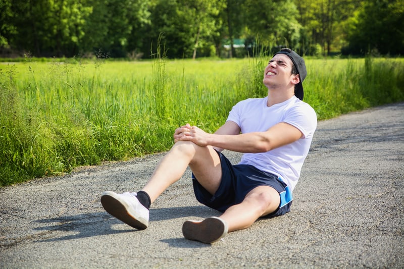 A man in a white t-shirt, navy blue shorts, and a backward black cap is sitting on the ground of a paved path, clutching his knee with a pained expression. He appears to be in a park or natural area with trees and grass in the background.