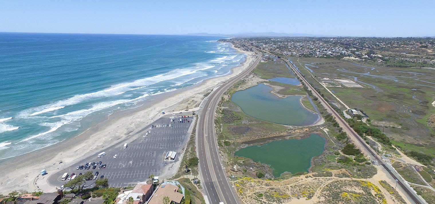 An aerial view captures a scenic coastline with a long, sandy beach on the left and a road running parallel to the shoreline. On the right, marshlands and small bodies of water can be seen, with a town in the distance and mountains on the horizon.